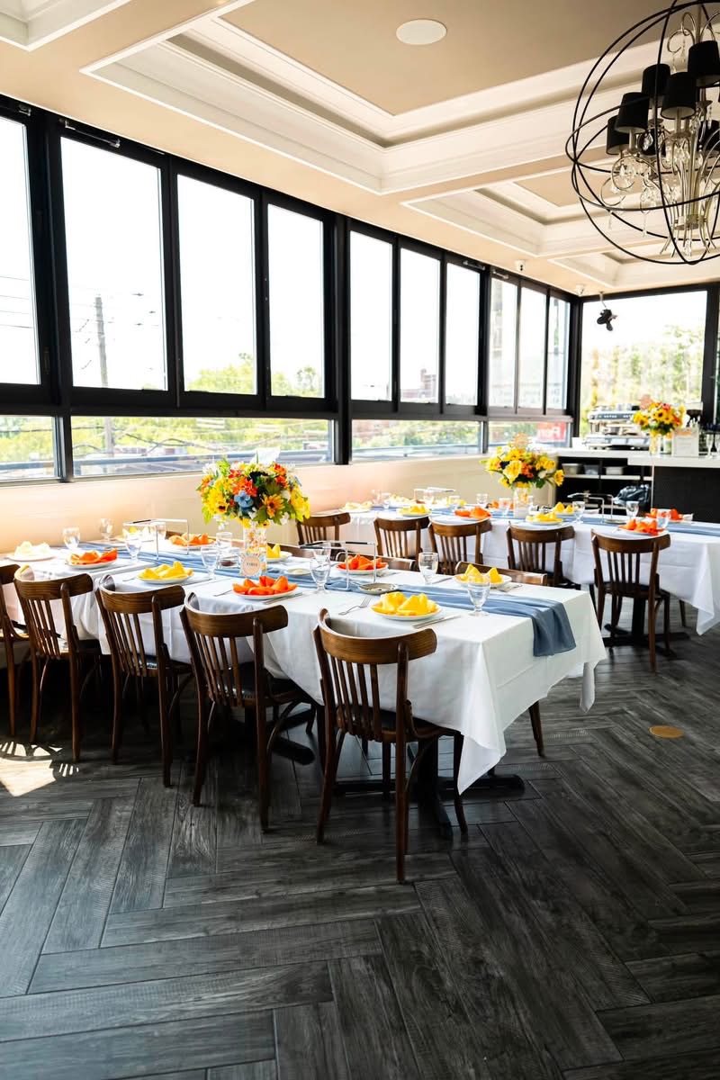 The Rooftop set for a celebration with white linens, blue runners and floral centerpieces beside the window wall